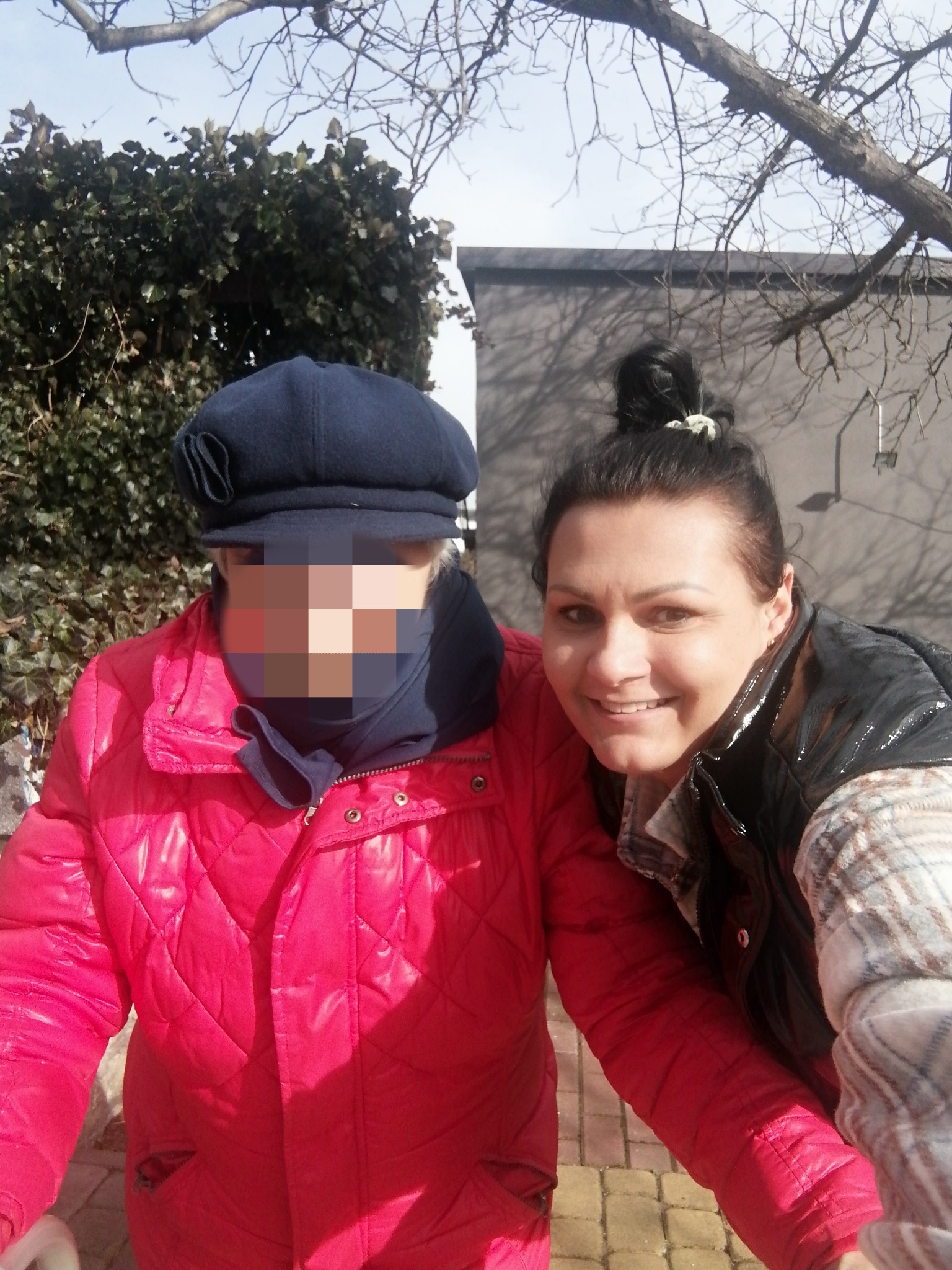 Kobieta w czarnej kurtce pozuje uśmiechnięta obok osoby starszej w czerwonej kurtce i czapce, której twarz jest zamazana, na zewnątrz w słoneczny dzień.