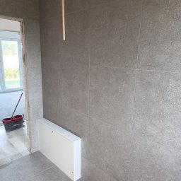 GENERAL-BUD Piotr Porczyński - Ściana i podłoga wyłożone szarymi płytkami imitującymi kamień, biała szafka rewizyjna przy podłodze, widoczne wiadro i mop w przejściu do kolejnego pomieszczenia.