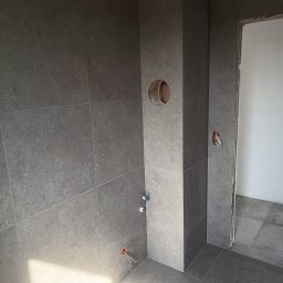 GENERAL-BUD Piotr Porczyński - Szare, matowe płytki wielkoformatowe na ścianach i podłodze łazienki w trakcie wykańczania, widoczne otwory na instalacje i niezamaskowane kable, surowe wykończenie ościeżnicy.