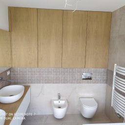 GENERAL-BUD Piotr Porczyński - Nowoczesna łazienka z beżowymi płytkami, drewnianymi szafkami nad umywalką i toaletą, widoczny bidet i grzejnik drabinkowy.