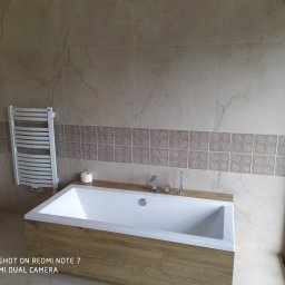 GENERAL-BUD Piotr Porczyński - Nowoczesna łazienka w beżowych odcieniach z wanną obudowaną drewnopodobną płytą, grzejnikiem drabinkowym i toaletą wiszącą.