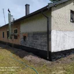 Dwukondygnacyjny budynek z cegły i pustaków, z widoczną izolacją fundamentów i fragmentem instalacji odgromowej na dachu, ukazany w słoneczny dzień z perspektywy trawnika.