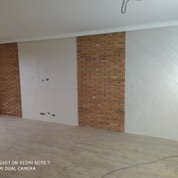 GENERAL-BUD Piotr Porczyński - Fragment pokoju z dekoracyjnym wykończeniem ścian: panele z cegły klinkierowej oraz ściany pokryte dekoracyjnym tynkiem w odcieniach bieli i szarości, widoczne gniazdka elektryczne i wykończona...