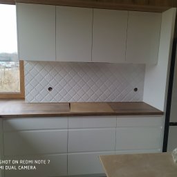 GENERAL-BUD Piotr Porczyński - Montaż białych szafek kuchennych z drewnianym blatem i dekoracyjną płytą nad blatem z otworami na gniazdka elektryczne, widok na okno i krajobraz za nim.