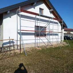 Biały dom jednorodzinny w trakcie prac elewacyjnych, z rozstawionymi rusztowaniami i widocznym fragmentem ogrodu.