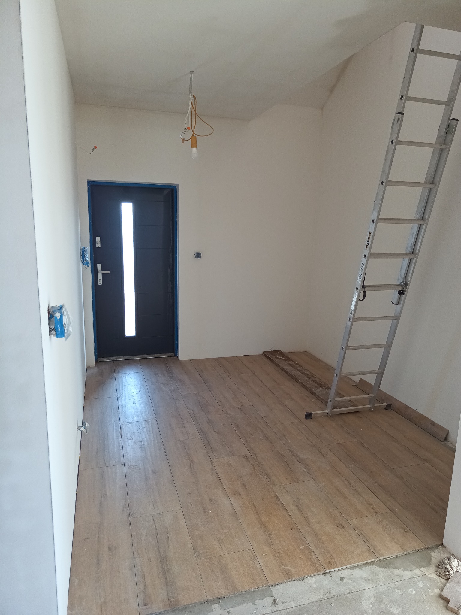 Wnętrze w trakcie prac wykończeniowych: położona podłoga z paneli imitujących drewno, białe ściany, czarne drzwi wejściowe z przeszkleniem, drabina oparta o ścianę, widoczne niedokończone prace...