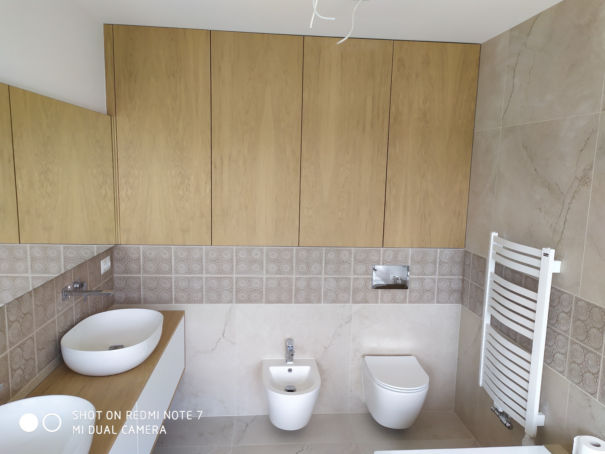 Nowoczesna łazienka z beżowymi płytkami, drewnianymi szafkami nad umywalką i toaletą, widoczny bidet i grzejnik drabinkowy.