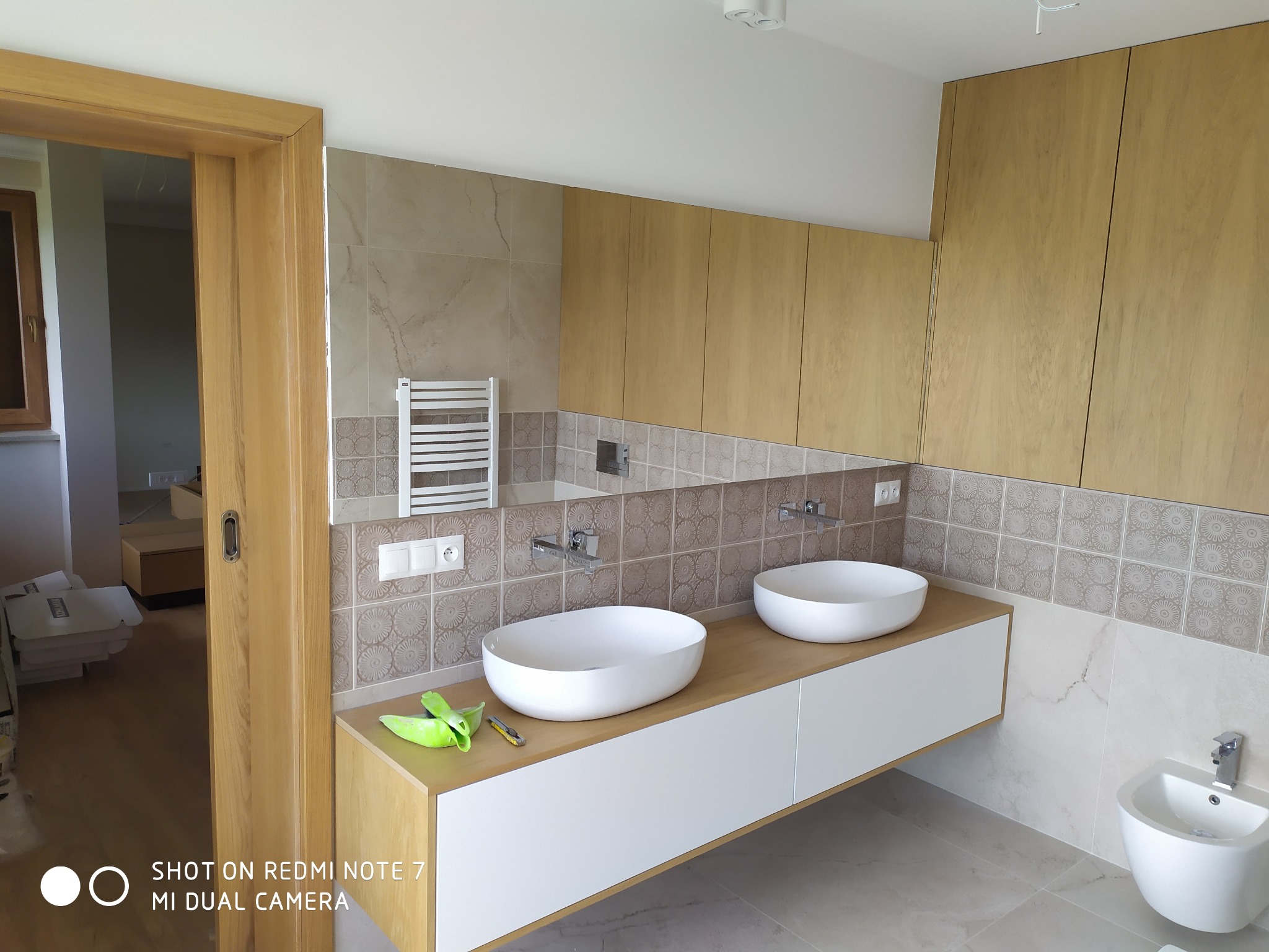 Nowoczesna łazienka z dwoma białymi umywalkami nablatowymi na drewnianym blacie, szafki wiszące z forniru, dekoracyjne płytki na ścianie, bidet i grzejnik drabinkowy.