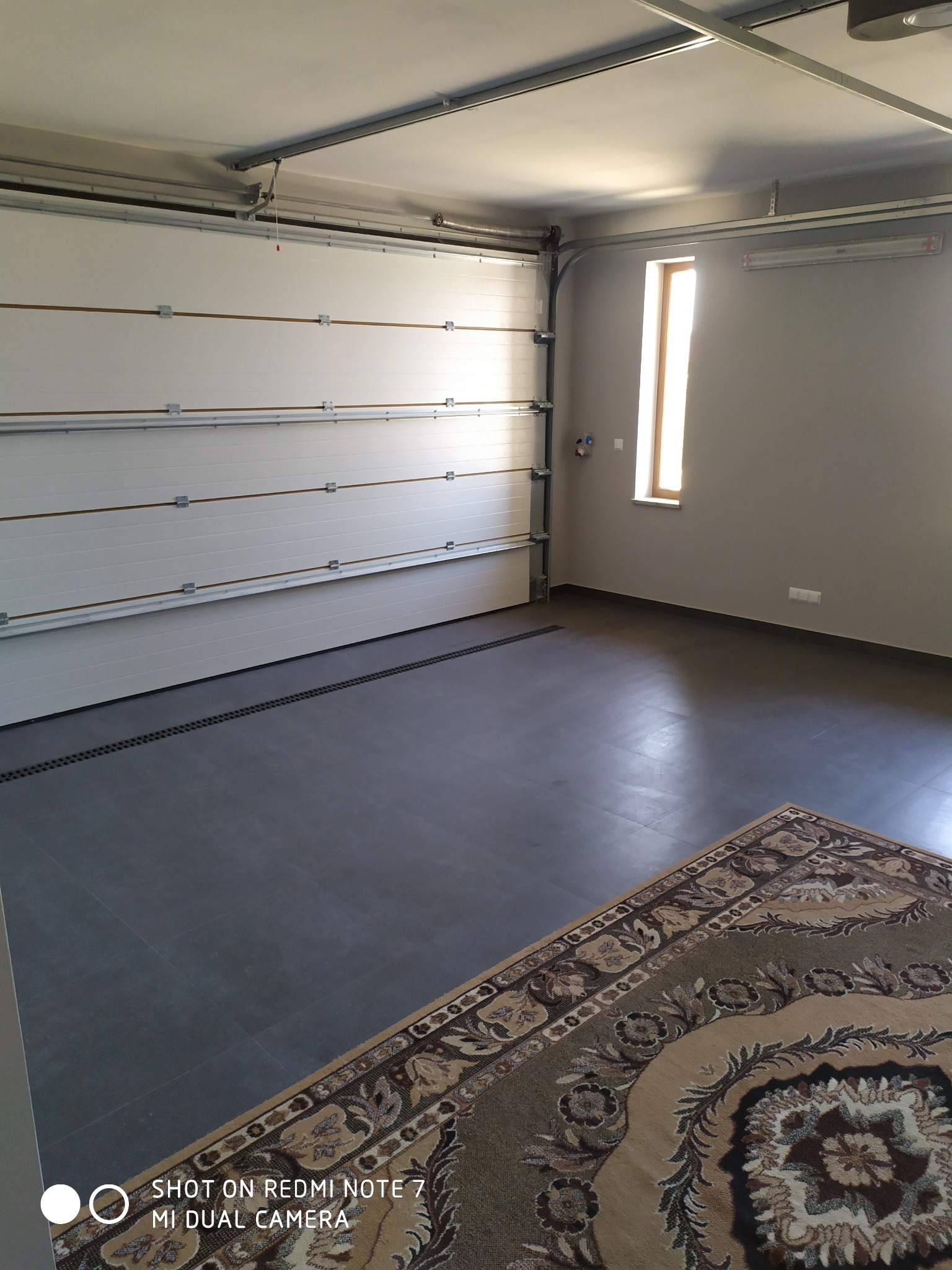 Wnętrze garażu z segmentową bramą garażową w kolorze białym, szarą podłogą z odpływem liniowym i beżowym dywanem z orientalnym wzorem w rogu, oświetlone naturalnym światłem z okna