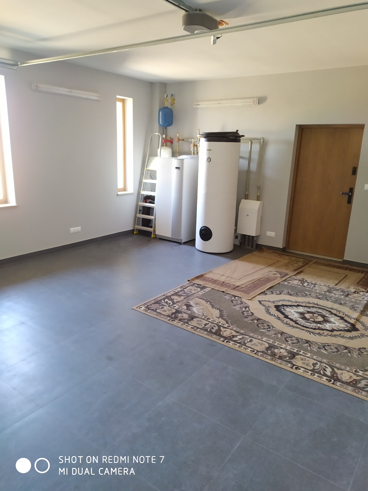 Szare płytki podłogowe w pomieszczeniu z zamontowanymi bojlerami i drabina oparta o ścianę, widoczne dwa dywany.