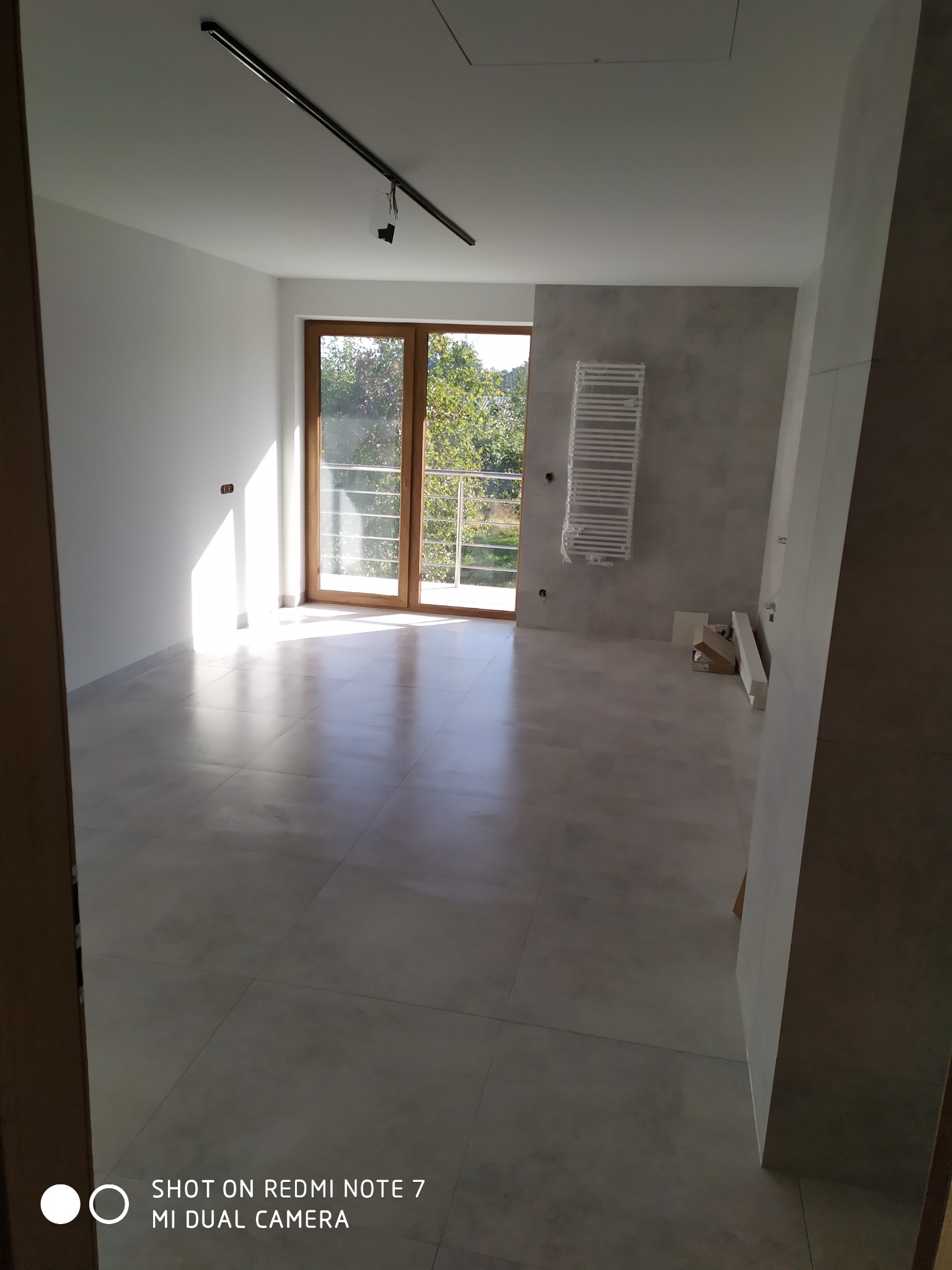 Jasne, przestronne wnętrze łazienki z dużym oknem balkonowym, grzejnikiem drabinkowym na szarej ścianie i szarą podłogą z płytek. Widoczne kartony i elementy wykończeniowe sugerują etap prac...