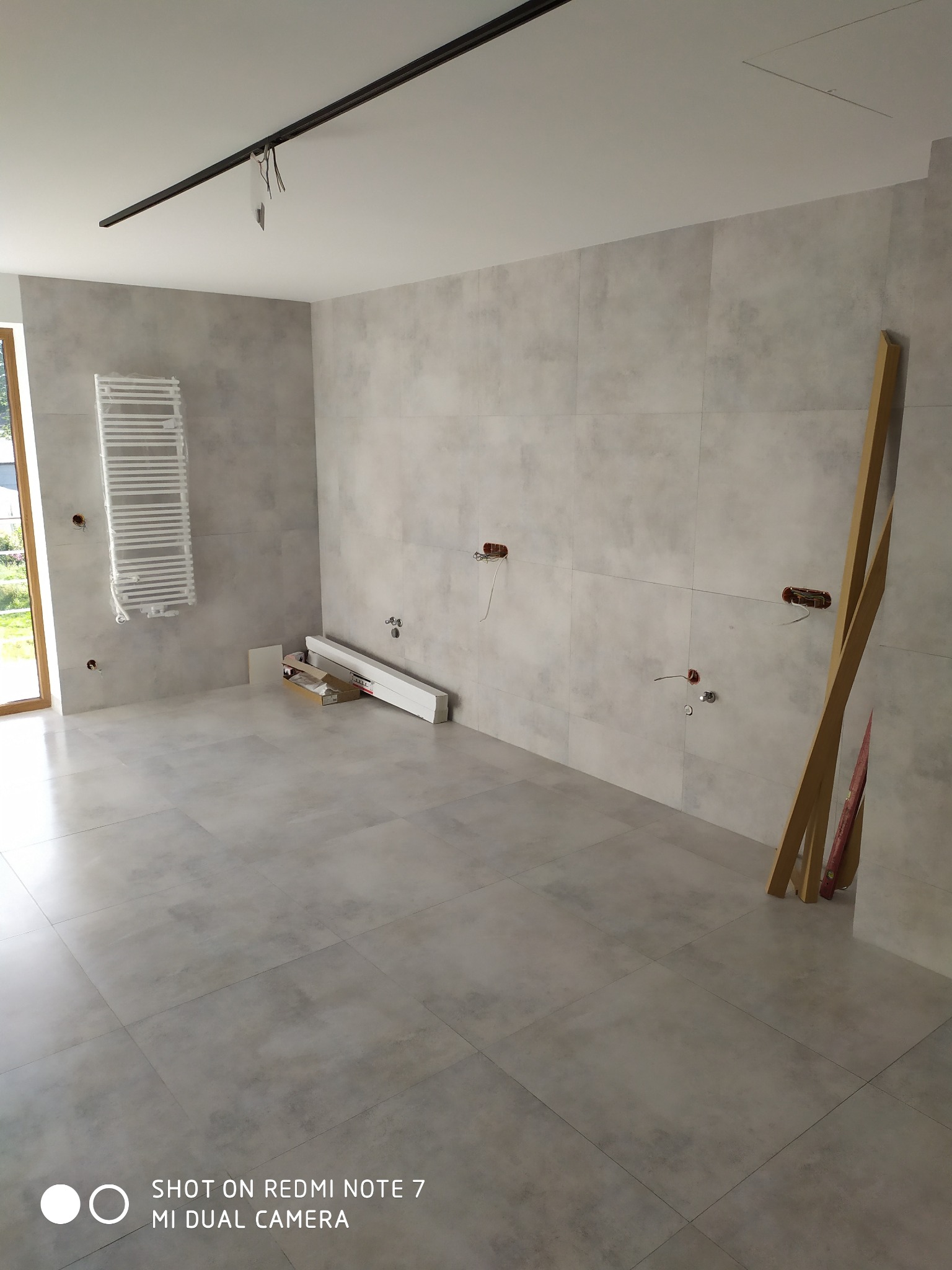 Wnętrze łazienki w trakcie prac wykończeniowych: szare płytki na ścianach i podłodze, grzejnik drabinkowy, przygotowane instalacje wodne i elektryczne, listwy przypodłogowe w opakowaniu, drewniane...