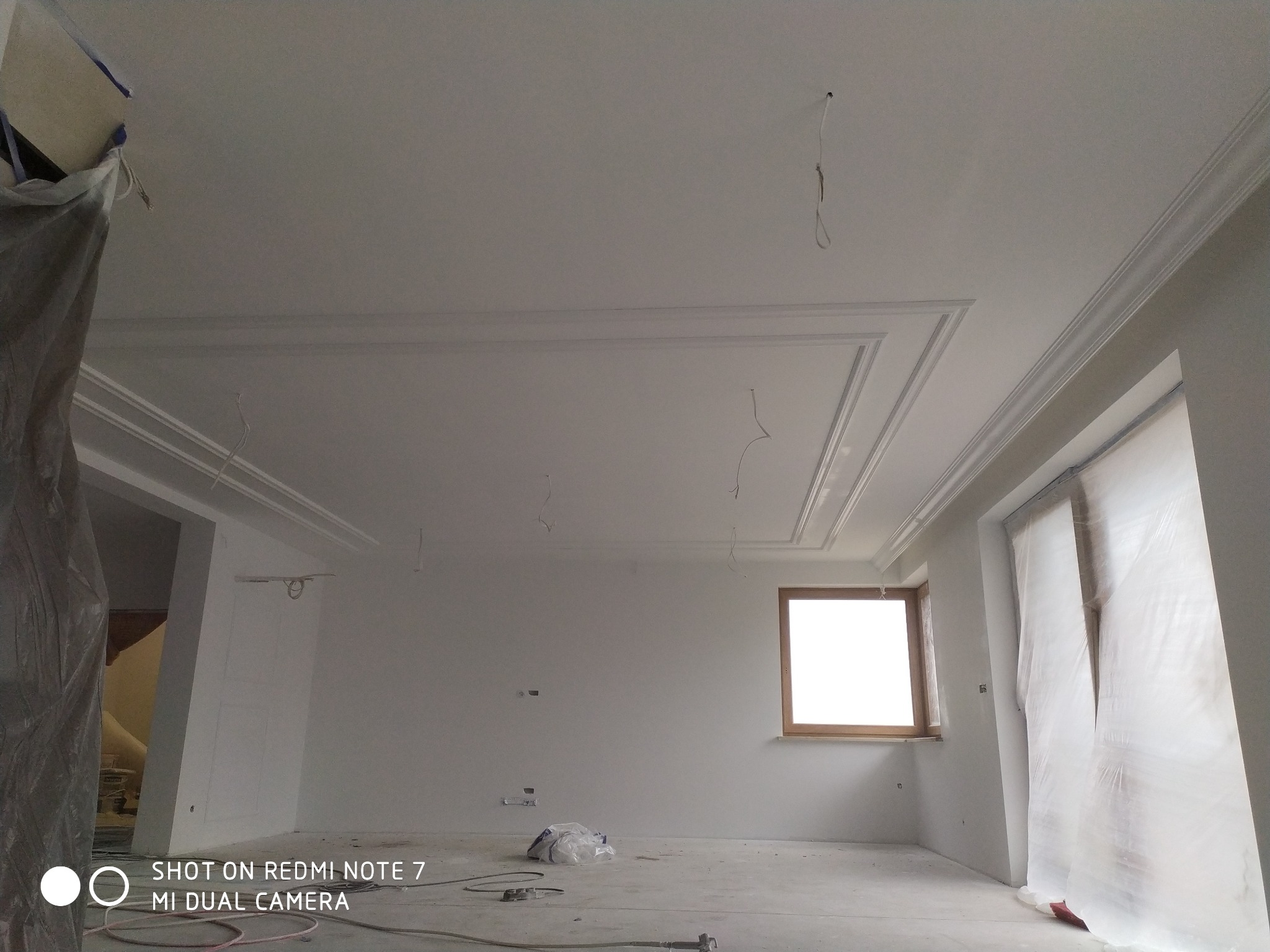 Wykończony sufit z dekoracyjnymi listwami w Dobroniu Małym, widoczne przygotowania do dalszych prac wykończeniowych, zakryte okno folią ochronną, kable wystające z sufitu.
