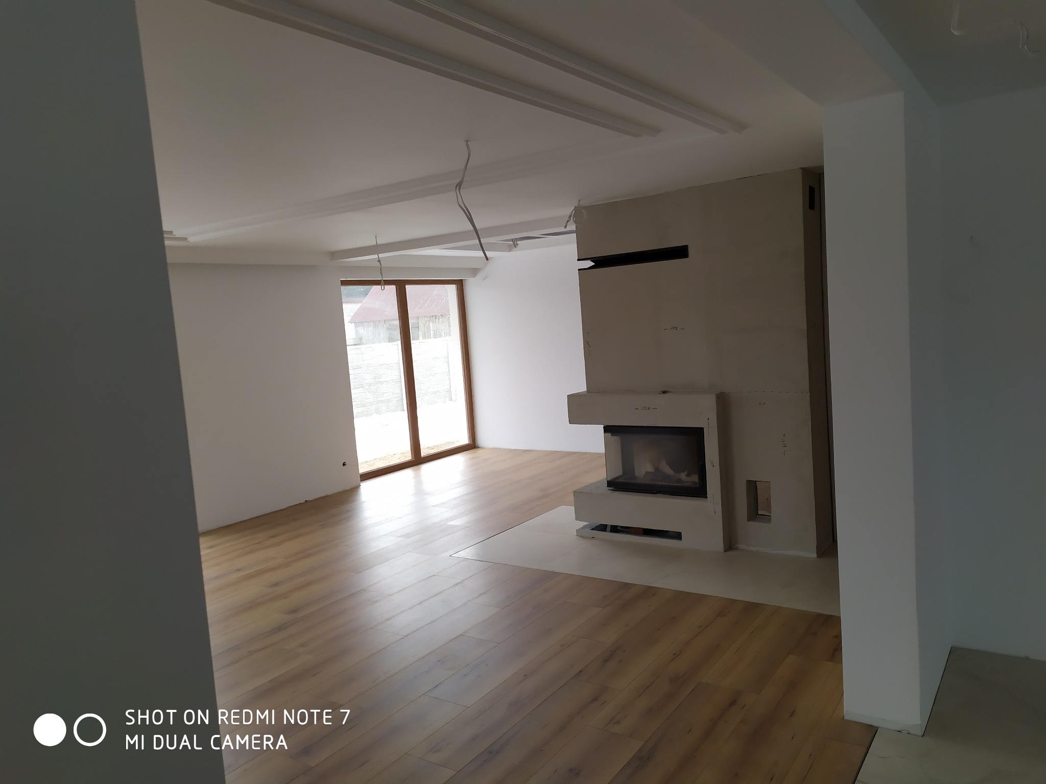 Salon z kominkiem w trakcie wykańczania, widoczne panele podłogowe, białe ściany, surowe wykończenie kominka i wystające kable z sufitu.