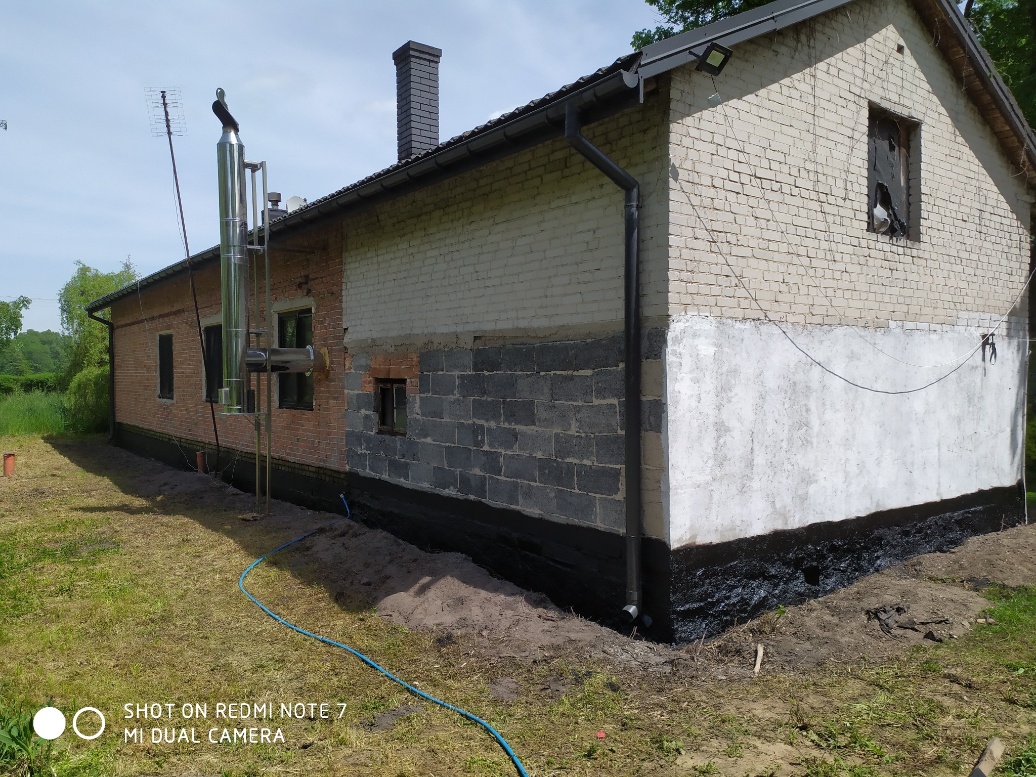 Dwukondygnacyjny budynek z cegły i pustaków, z widoczną izolacją fundamentów i fragmentem instalacji odgromowej na dachu, ukazany w słoneczny dzień z perspektywy trawnika.