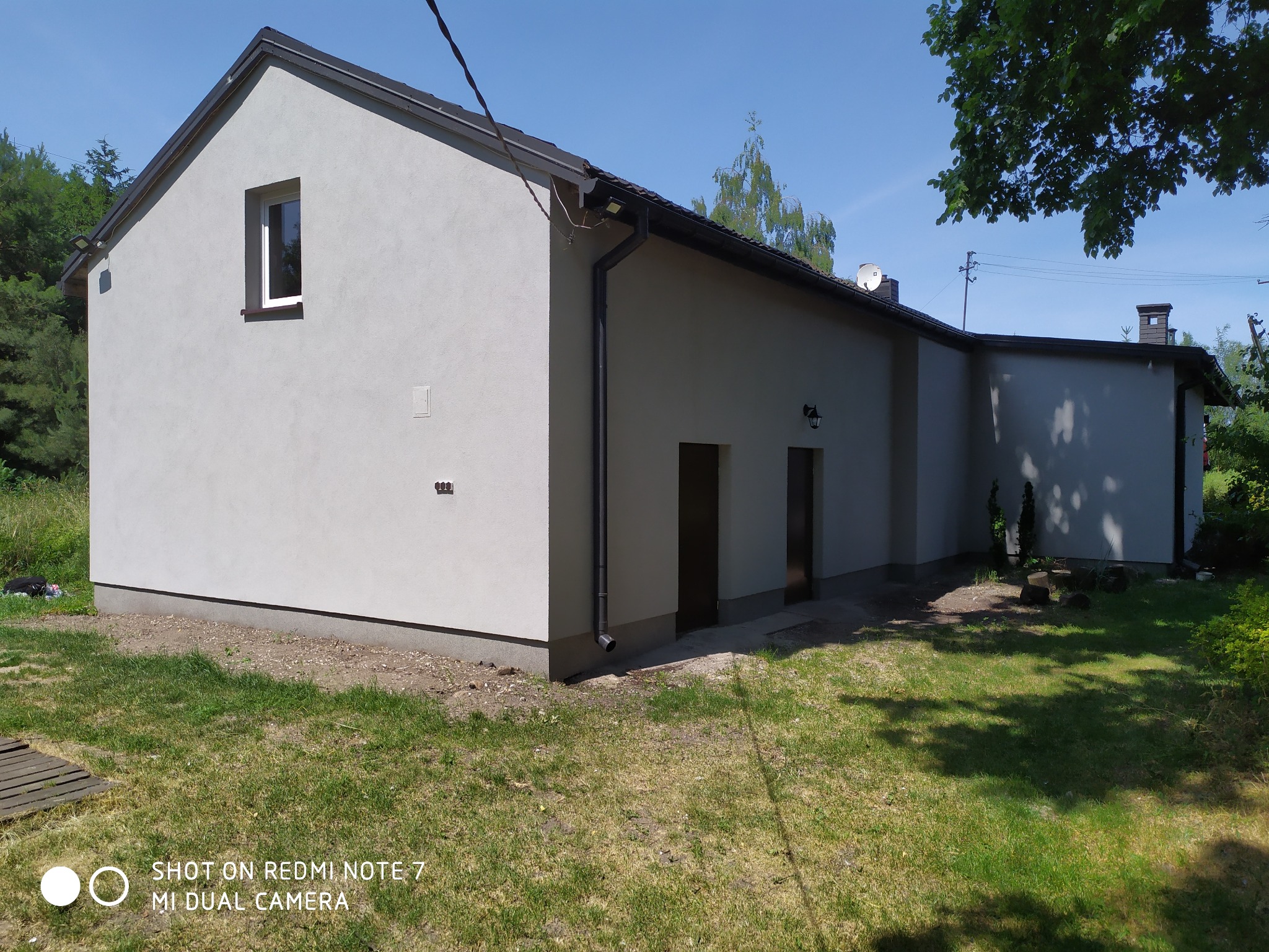 Świeżo otynkowany, jasnoszary budynek z ciemną dachówką i białymi ramami okiennymi, widoczny z zewnątrz w słoneczny dzień, z fragmentem ogrodu i drzew w tle.