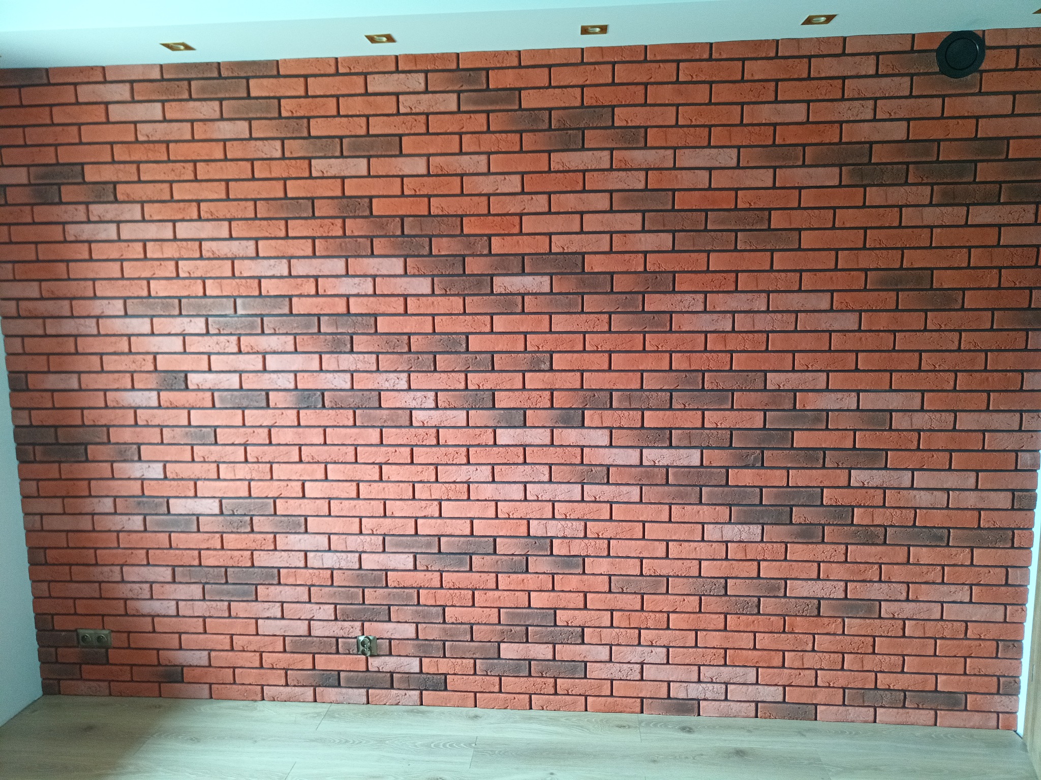 Ściana wyłożona dekoracyjną cegłą w odcieniach czerwieni i brązu, widoczne gniazdka elektryczne i wentylacja, jasna podłoga i sufit z oświetleniem punktowym.