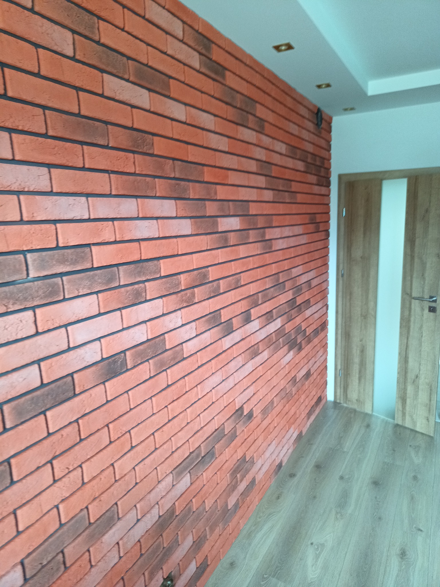 Ściana z cegły klinkierowej w odcieniach czerwieni i brązu, z widoczną strukturą i fugami, przylegająca do drewnianej podłogi i drzwi z przeszkleniem w nowoczesnym wnętrzu.