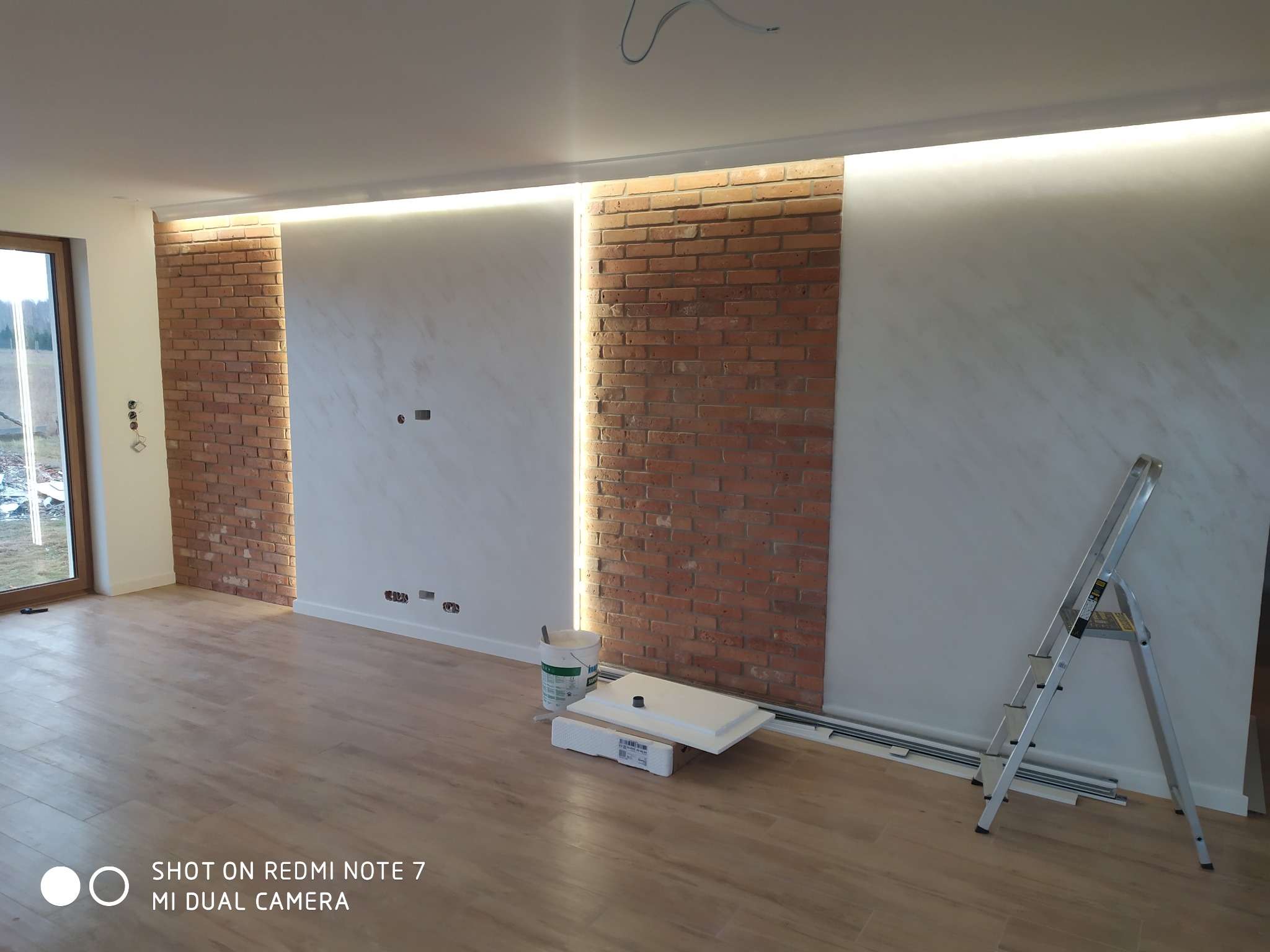 Wykończone wnętrze pokoju z dekoracyjną ścianą z cegły, oświetleniem LED w górnej części ściany, pomalowanymi ścianami z efektem marmuru, drewnopodobną podłogą, drabina i akcesoria malarskie...