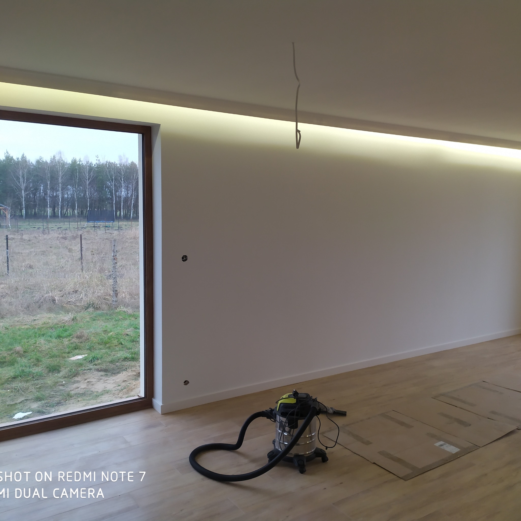 Wnętrze pokoju po pracach wykończeniowych: białe ściany z widocznymi puszkami elektrycznymi, jasna podłoga zabezpieczona kartonem, okno z widokiem na zewnątrz, oraz odkurzacz przemysłowy stojący...