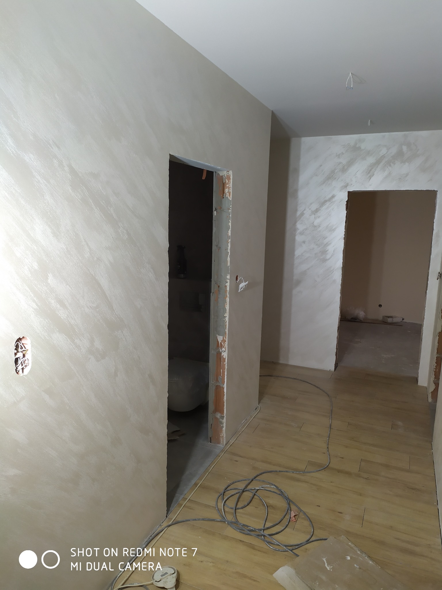 Wnętrze mieszkania w trakcie prac wykończeniowych, ściany pokryte dekoracyjną, perłową farbą, widoczne niedokończone ościeżnice, kabel elektryczny na podłodze z paneli.