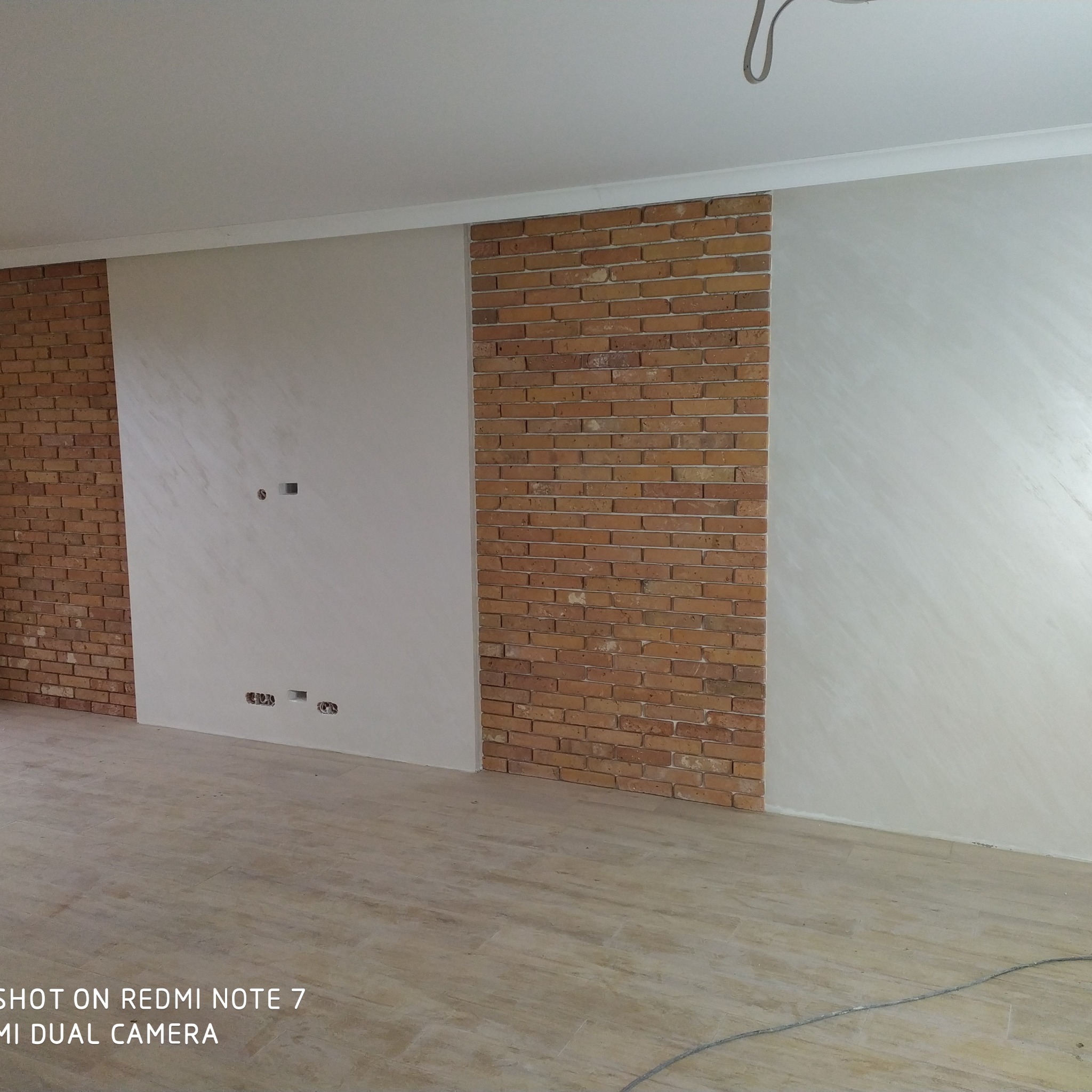 Fragment pokoju z dekoracyjnym wykończeniem ścian: panele z cegły klinkierowej oraz ściany pokryte dekoracyjnym tynkiem w odcieniach bieli i szarości, widoczne gniazdka elektryczne i wykończona...