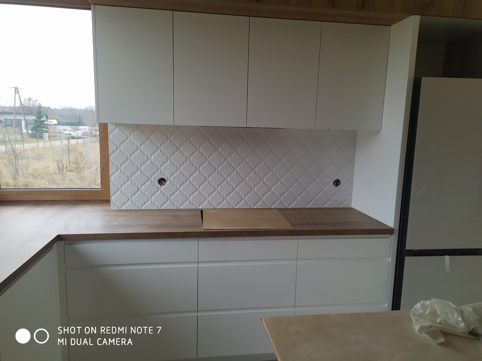 Montaż białych szafek kuchennych z drewnianym blatem i dekoracyjną płytą nad blatem z otworami na gniazdka elektryczne, widok na okno i krajobraz za nim.