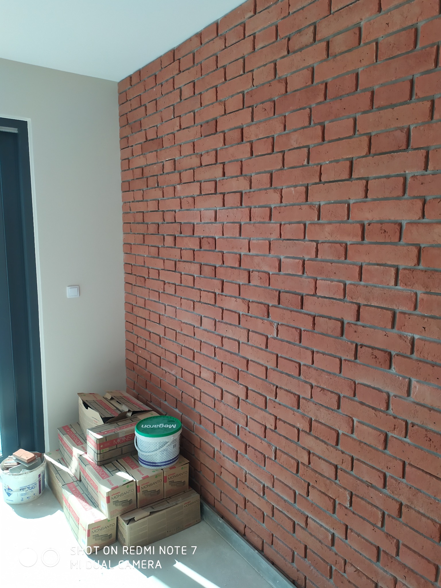 Ściana z cegieł dekoracyjnych w Dobroniu Małym, z widocznymi materiałami budowlanymi w kartonach i wiaderkach, czekająca na dalsze prace wykończeniowe.