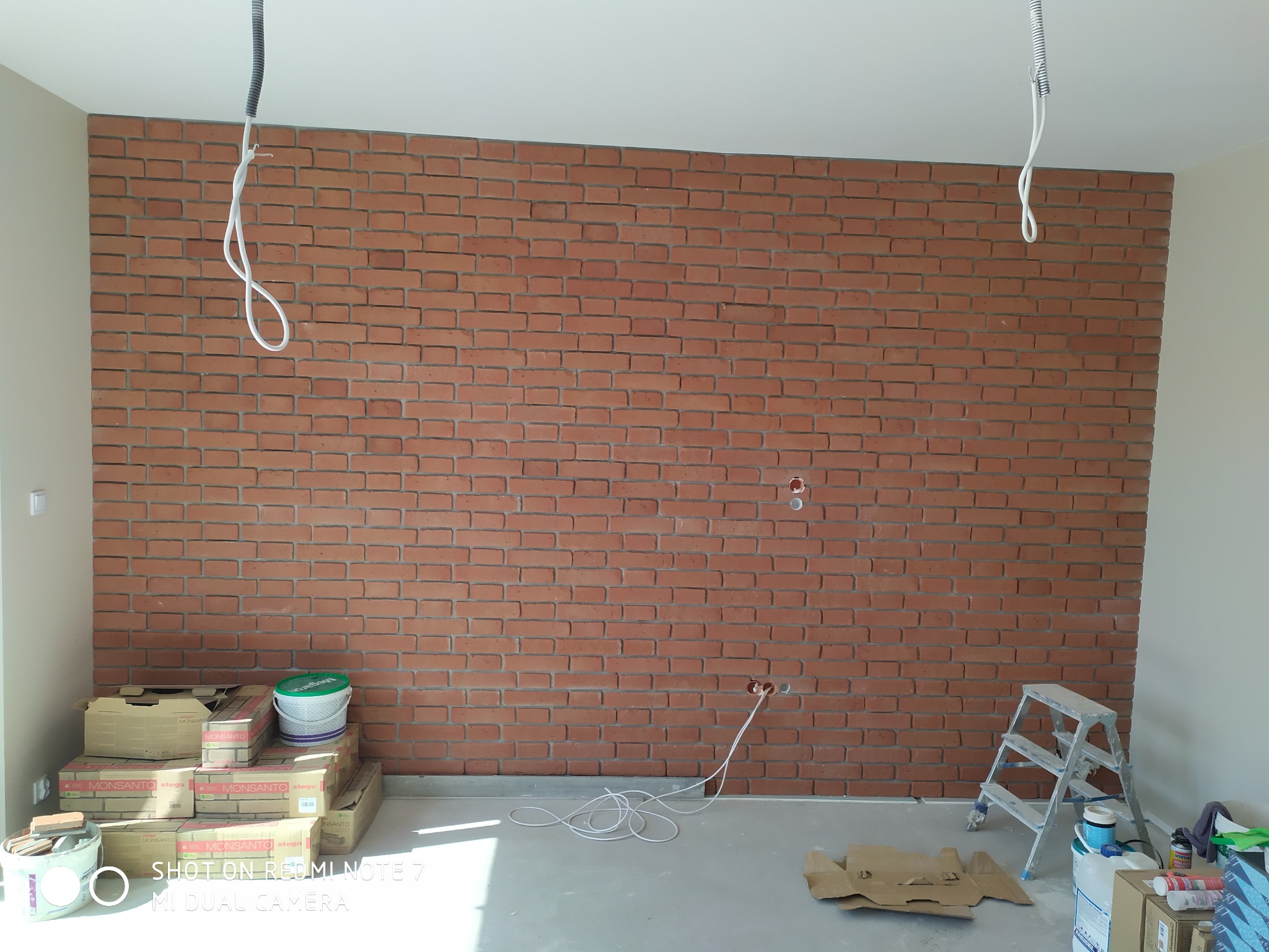 Ściana wyłożona czerwoną cegłą dekoracyjną w trakcie prac wykończeniowych, widoczne puszki elektryczne, kable, drabina, opakowania cegieł i materiały budowlane na podłodze.