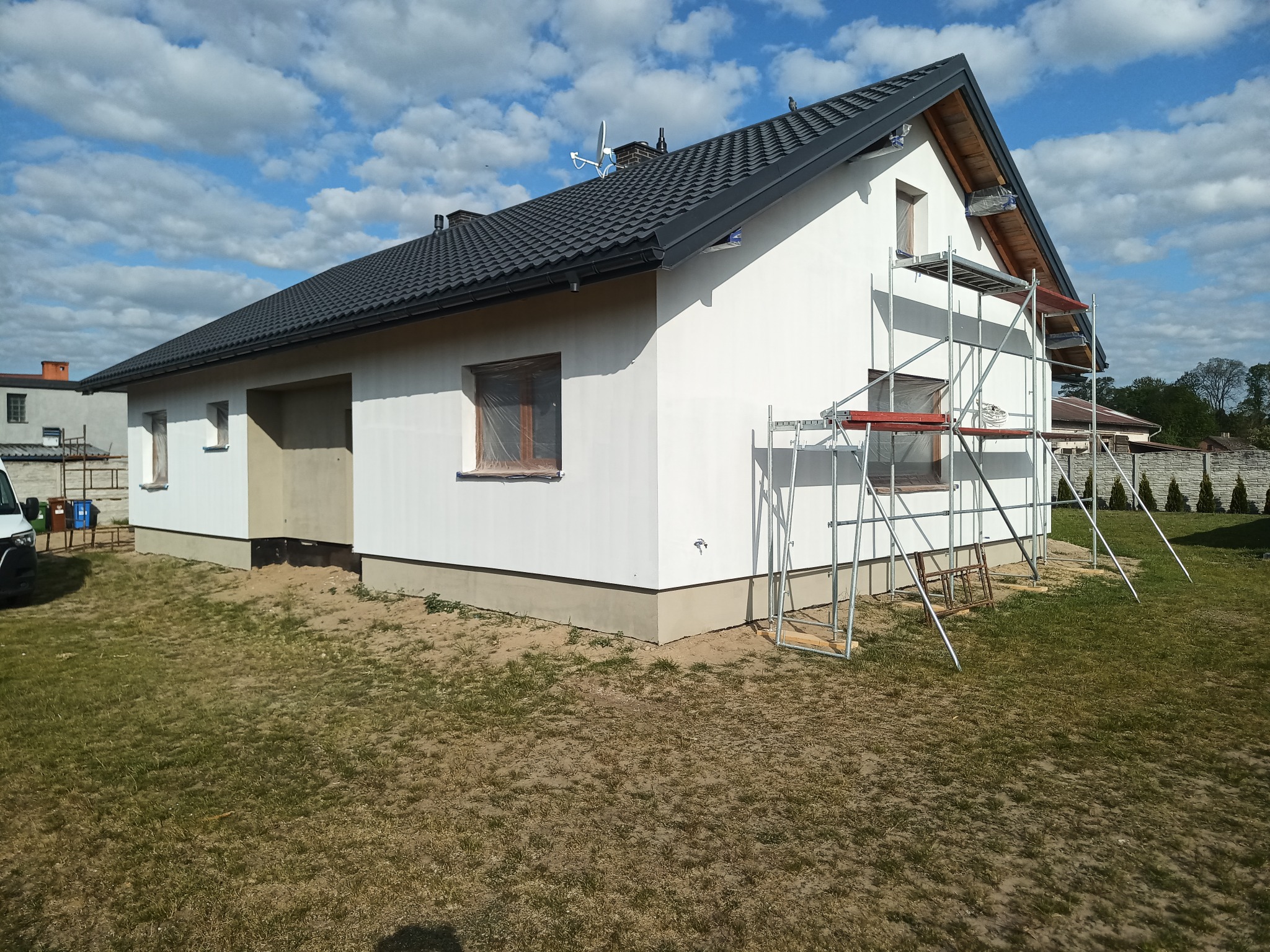 Nowy, biały dom jednorodzinny z ciemną dachówką, częściowo osłonięte okna, rusztowania przy ścianie szczytowej, błękitne niebo w tle.