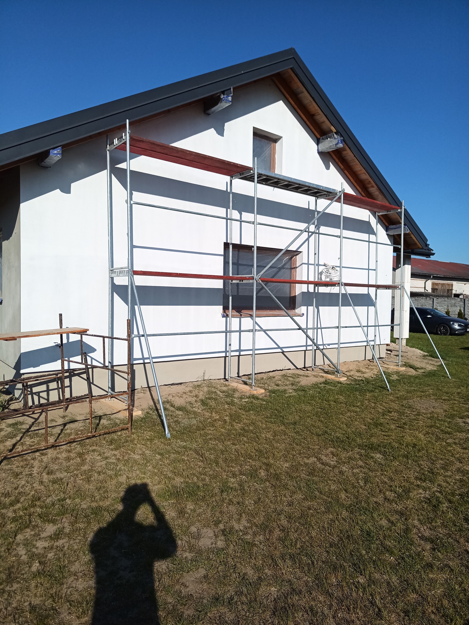 Biały dom jednorodzinny w trakcie prac elewacyjnych, z rozstawionymi rusztowaniami i widocznym fragmentem ogrodu.