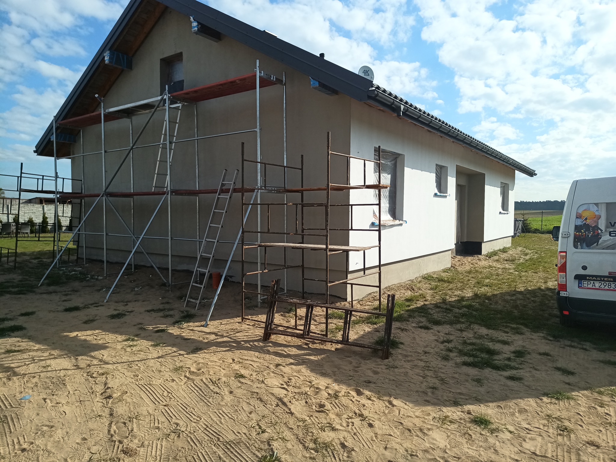 Ocieplanie domu jednorodzinnego w trakcie realizacji. Część elewacji otynkowana, część biała. Widoczne rusztowania i piasek wokół budynku. Błękitne niebo.