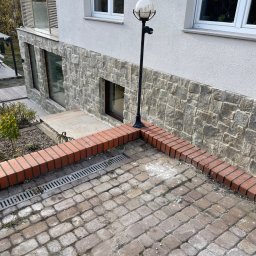 BEAR BUD MICHAŁ NIEDŹWIEDZKI - Elewacja domu z kamienia, czerwona ceglana obwódka rabaty, kostka brukowa i lampa ogrodowa z okrągłym kloszem.