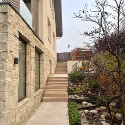 BEAR BUD MICHAŁ NIEDŹWIEDZKI - Elewacja domu z kamienia z drewnianymi schodami prowadzącymi do góry, widoczny kontener budowlany w tle