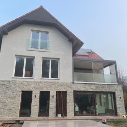 BEAR BUD MICHAŁ NIEDŹWIEDZKI - Nowoczesny dom jednorodzinny z elewacją z kamienia naturalnego, tarasem wyłożonym płytami i zabudowanym balkonem z panelami przeciwsłonecznymi na dachu.