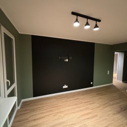 BEAR BUD MICHAŁ NIEDŹWIEDZKI - Pusty pokój z panelami podłogowymi, ścianą pomalowaną na czarno z uchwytem na telewizor i zielonymi ścianami bocznymi, oświetlony trzypunktową lampą sufitową.