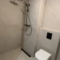 Nowoczesna łazienka z beżowymi płytkami imitującymi kamień, kabina prysznicowa z deszczownicą i odpływem liniowym oraz podwieszana toaleta z czarnym przyciskiem spłukującym.