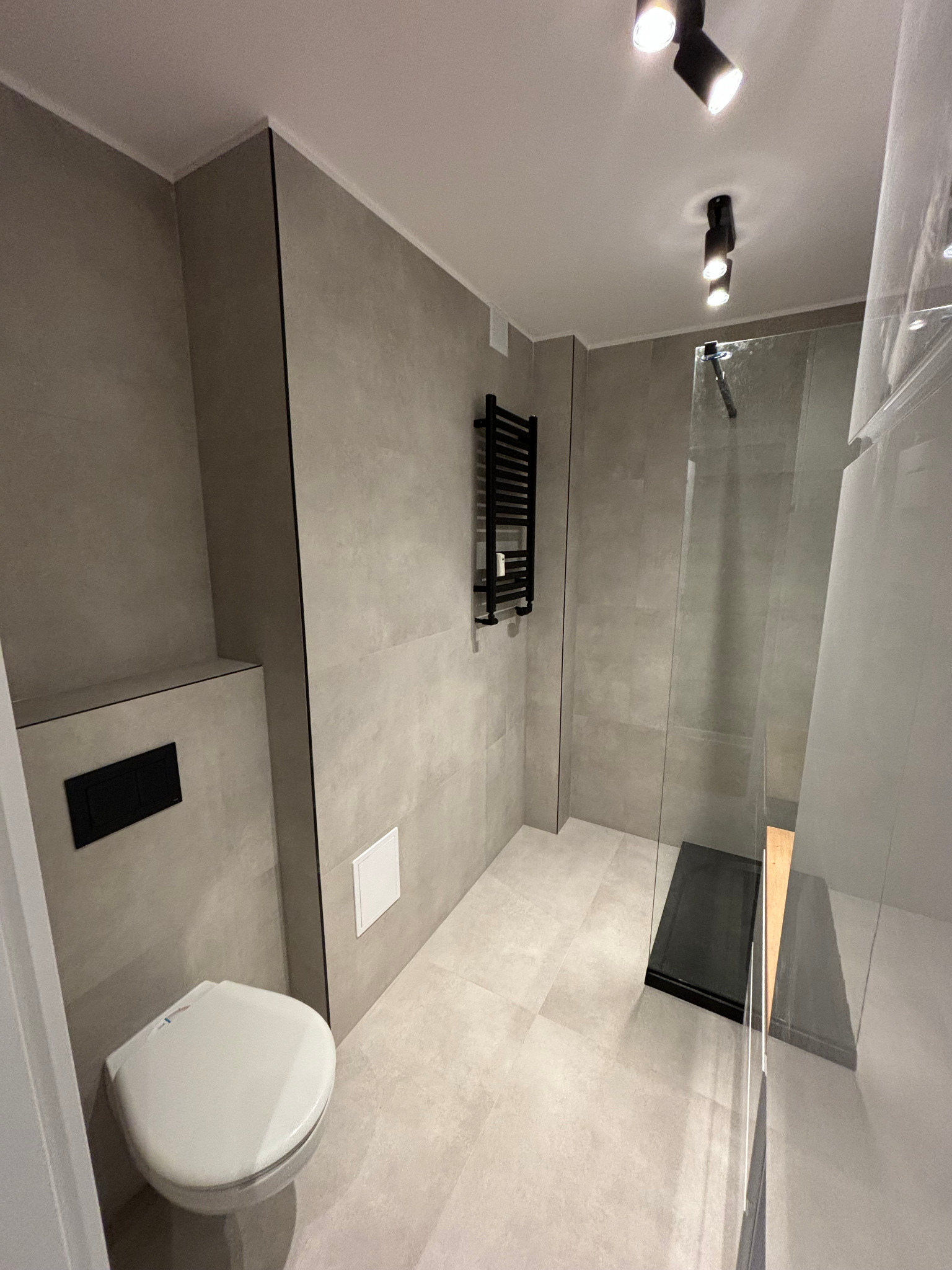 Nowoczesna łazienka z szarymi płytkami, wiszącą toaletą z czarnym przyciskiem, czarnym grzejnikiem drabinkowym i kabiną prysznicową typu walk-in z czarnym brodzikiem.