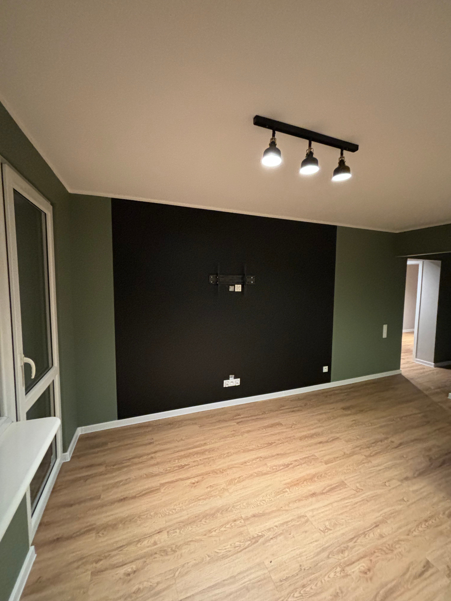 Pusty pokój z panelami podłogowymi, ścianą pomalowaną na czarno z uchwytem na telewizor i zielonymi ścianami bocznymi, oświetlony trzypunktową lampą sufitową.