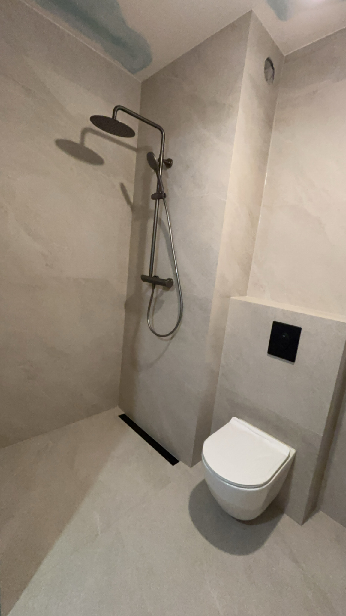 Nowoczesna łazienka z beżowymi płytkami imitującymi kamień, kabina prysznicowa z deszczownicą i odpływem liniowym oraz podwieszana toaleta z czarnym przyciskiem spłukującym.