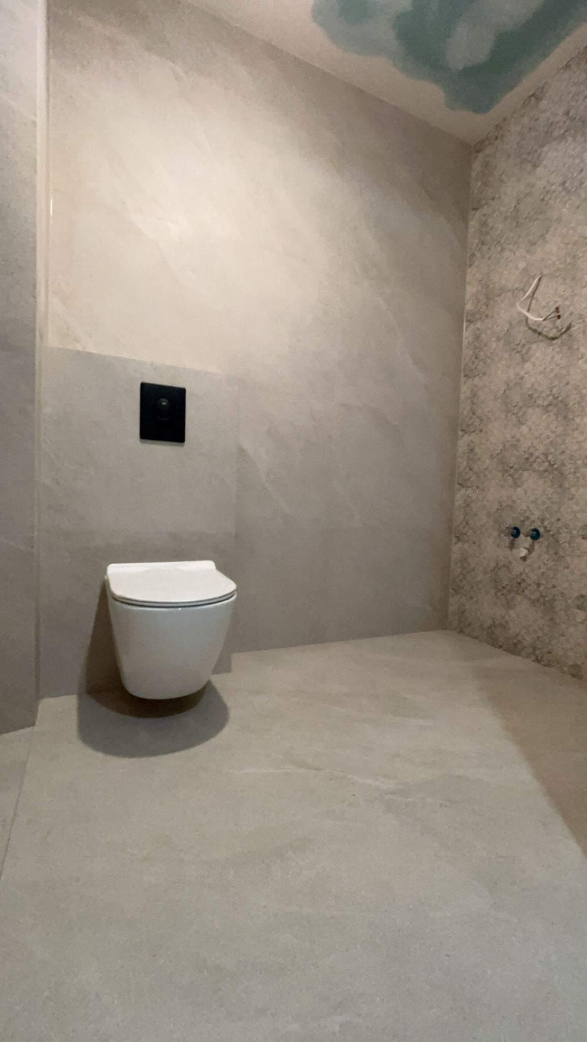 Świeżo wyremontowana łazienka z podwieszaną toaletą, szare płytki imitujące kamień na ścianach i podłodze, oraz dekoracyjna mozaika na jednej ze ścian, z widocznymi instalacjami wodnymi.