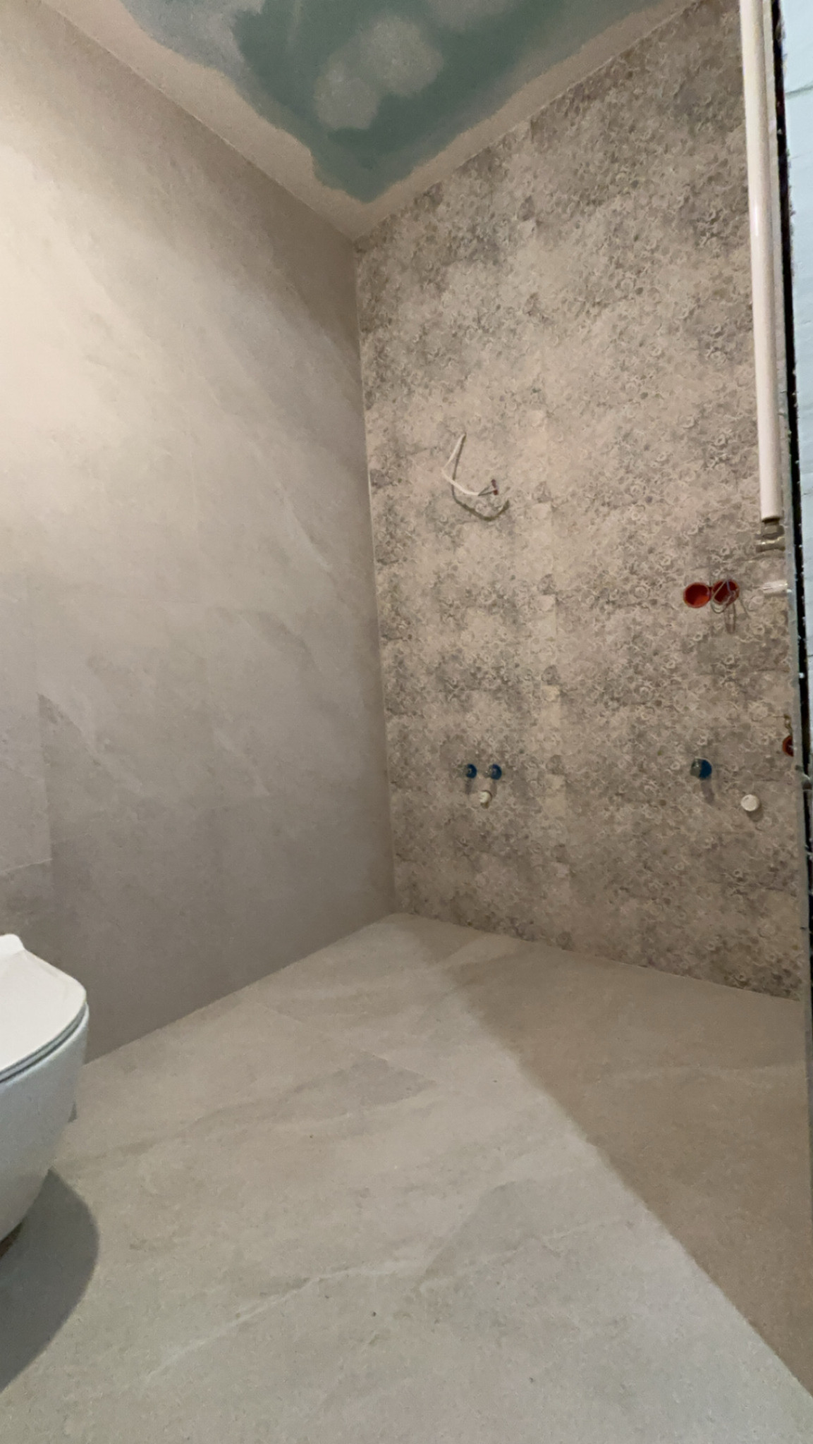 Niedokończona łazienka z szarymi płytkami o wzorzystej fakturze na ścianie, widoczne wyprowadzenia instalacji wodnej i elektrycznej, sufit częściowo pokryty niebieskim gruntem, w rogu widoczny...