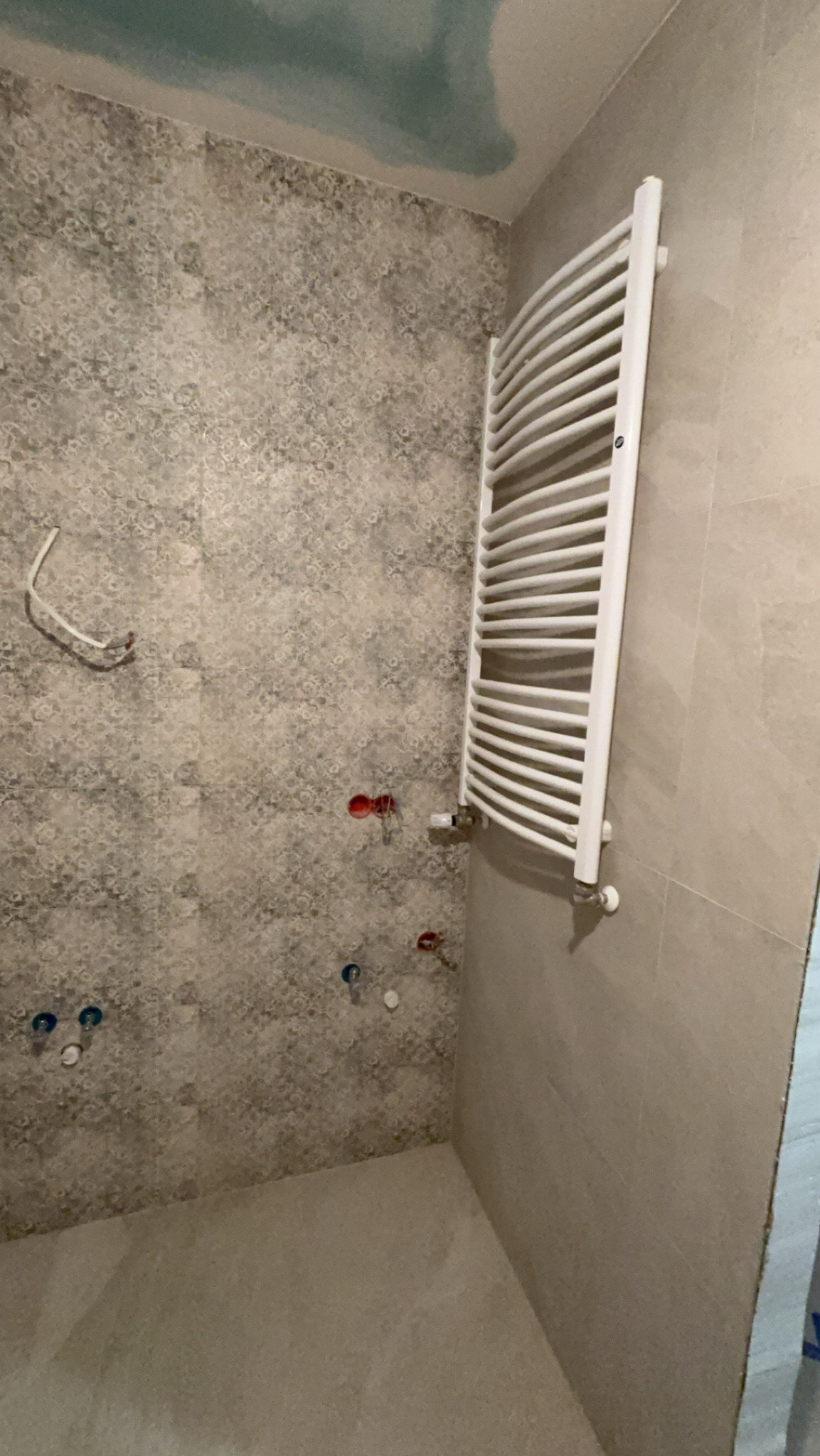 Ściana łazienki wyłożona wzorzystymi płytkami, z zamontowanym białym grzejnikiem drabinkowym i wystającymi rurami instalacji wodnej przed podłączeniem armatury.