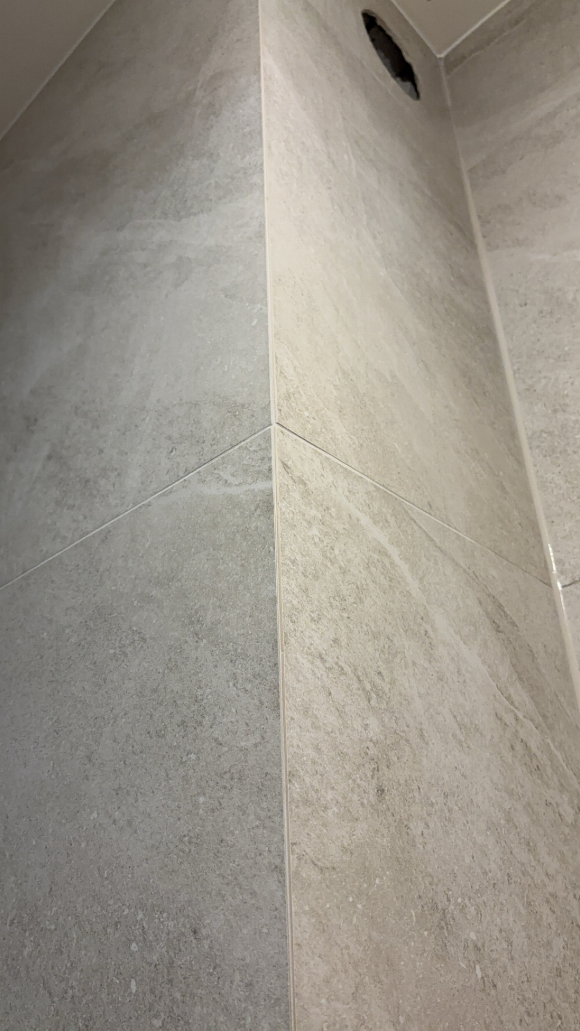 Szare i beżowe płytki imitujące kamień, ułożone w rogu łazienki z widocznym otworem wentylacyjnym w suficie.