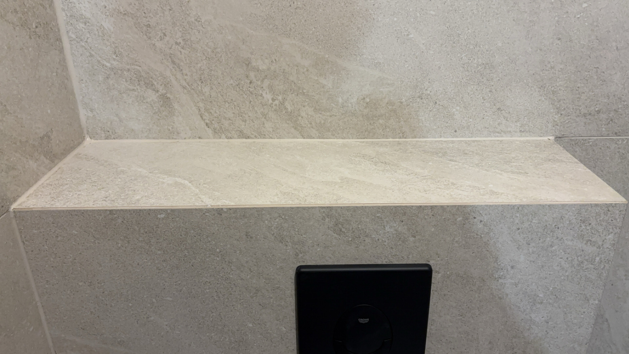 Wykończona wnęka w łazience wyłożona szarobeżowymi płytkami imitującymi kamień, z zamontowanym czarnym przyciskiem spłuczki Grohe.