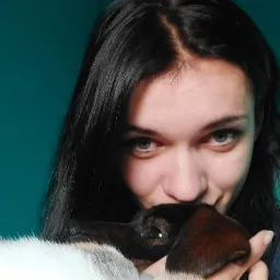 Portret kobiety z długimi, ciemnymi włosami, przytulającej trójkolorowego kota na tle turkusowej ściany.