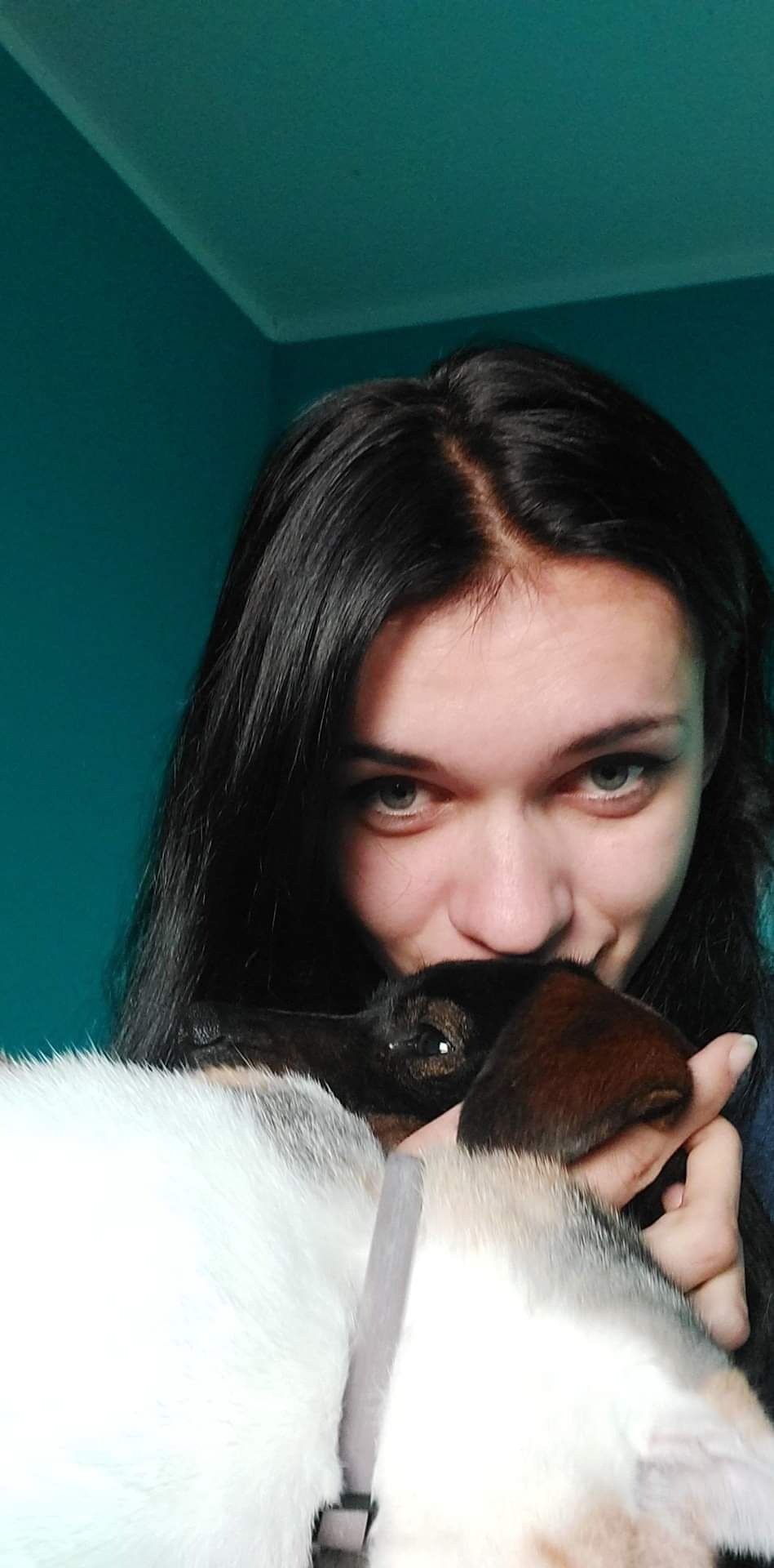 Portret kobiety z długimi, ciemnymi włosami, przytulającej trójkolorowego kota na tle turkusowej ściany.