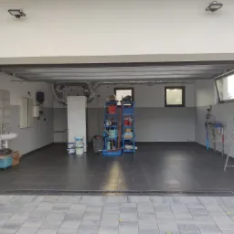 Wykończony garaż z szarymi płytkami podłogowymi i ściennymi, widoczna umywalka, regał z narzędziami, drabina, instalacja wentylacyjna pod sufitem, okna oraz lampa sufitowa.