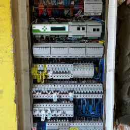 Otwarta skrzynka elektryczna z widocznymi zabezpieczeniami, przewodami i sterownikami Smart Home, zamontowana w surowym murze z fragmentami żółtej ściany po lewej stronie.