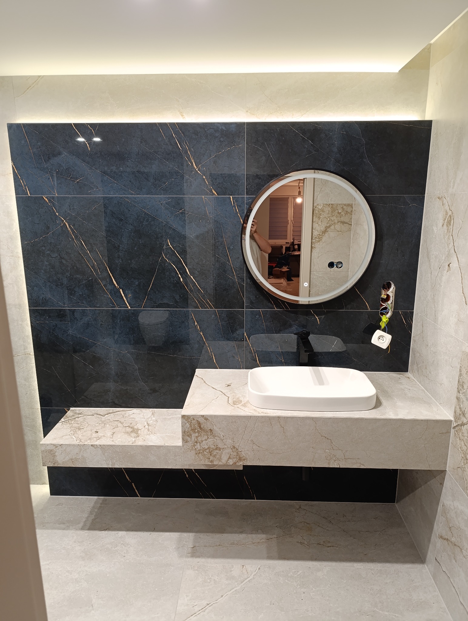 Elegancka łazienka z czarnymi płytkami z złotymi żyłami, okrągłym lustrem z podświetleniem i białą umywalką nablatową na marmurowym blacie.